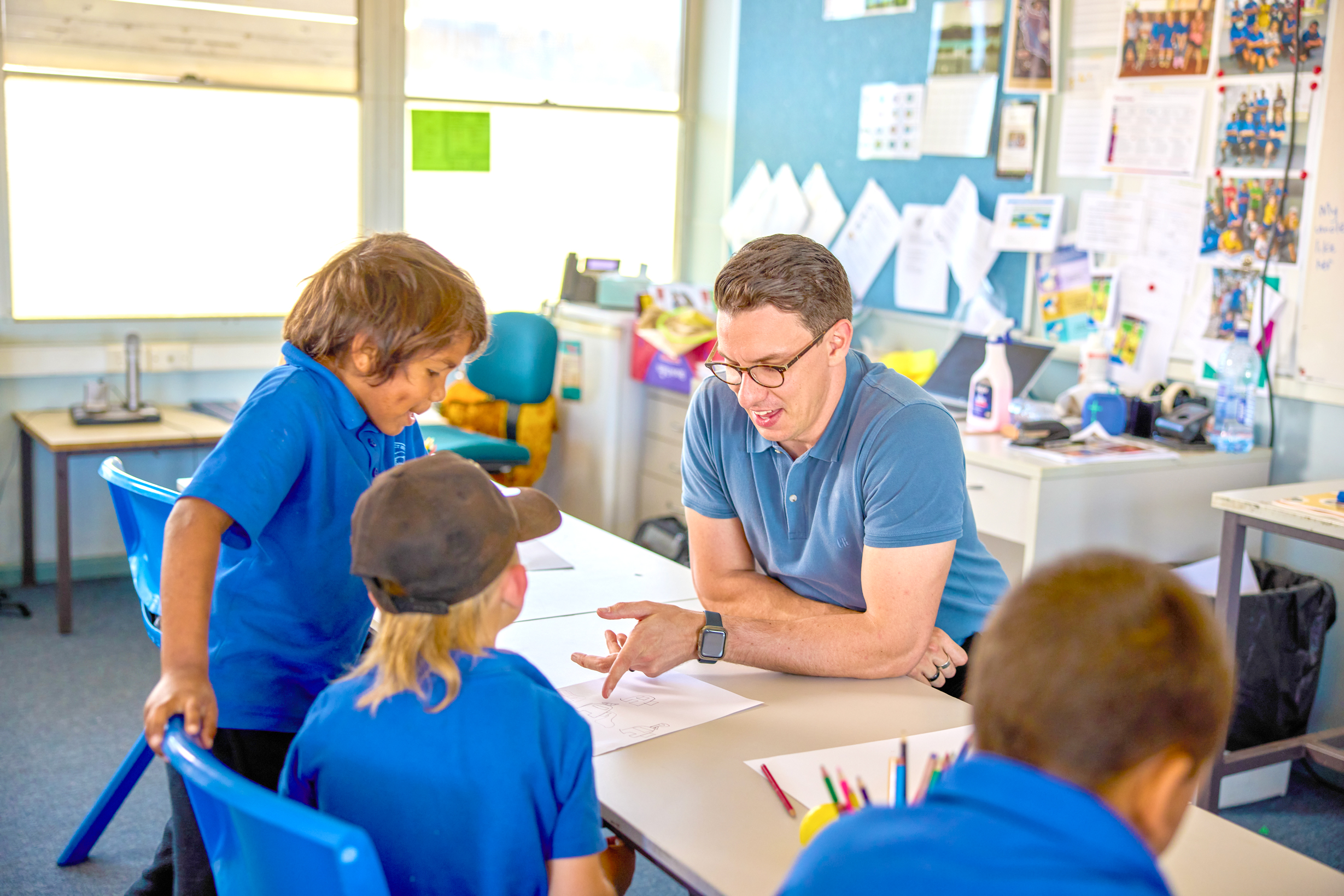 A teacher engages with young students, offering guidance at a classroom table.