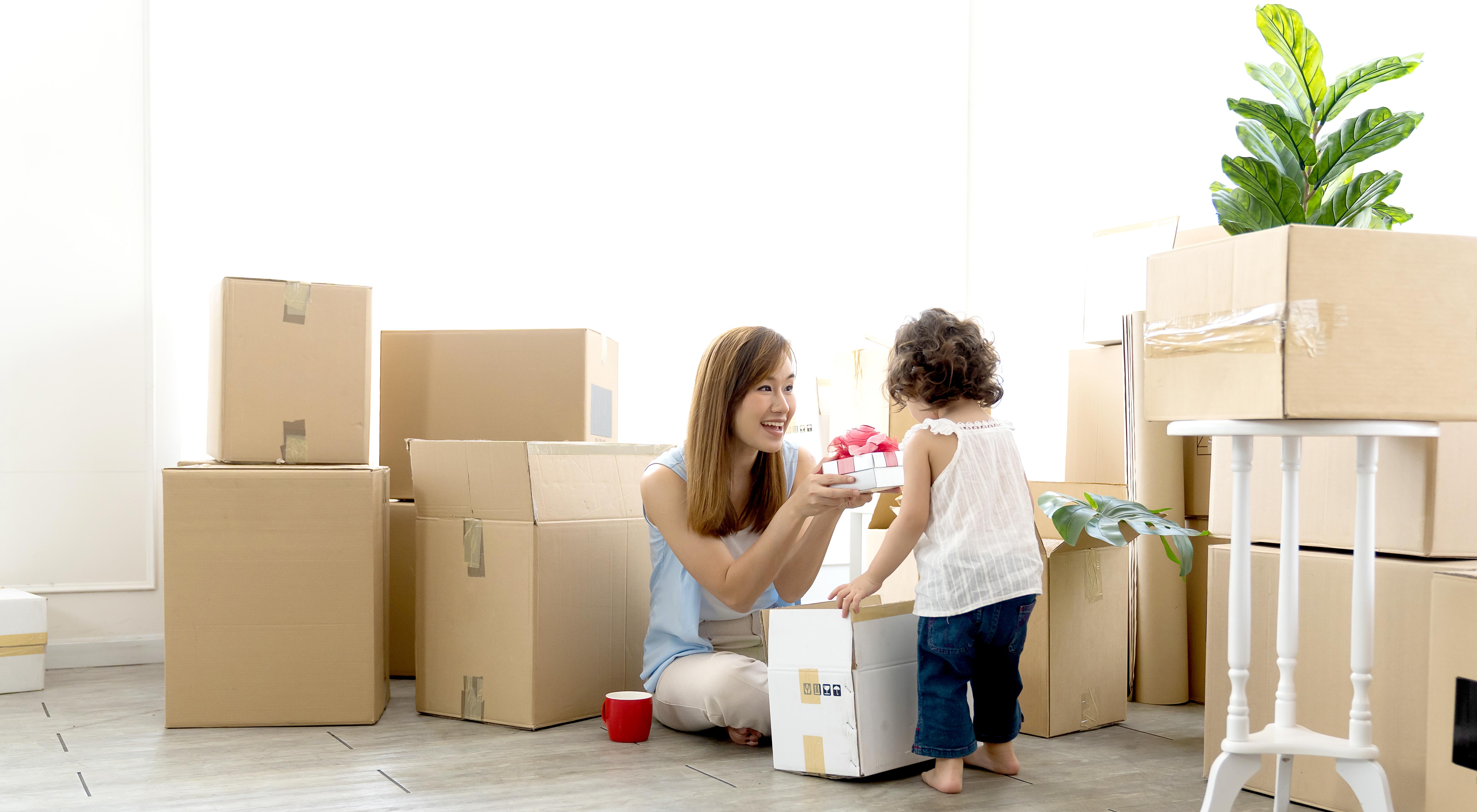 The image shows a woman and a child in a room filled with cardboard boxes, likely in the process of moving. The woman is kneeling on the floor with a warm smile, holding a gift box with a pink ribbon, which she is presenting to the child. The child, dressed in a white top and jeans, is standing in front of the woman, focused on the gift.