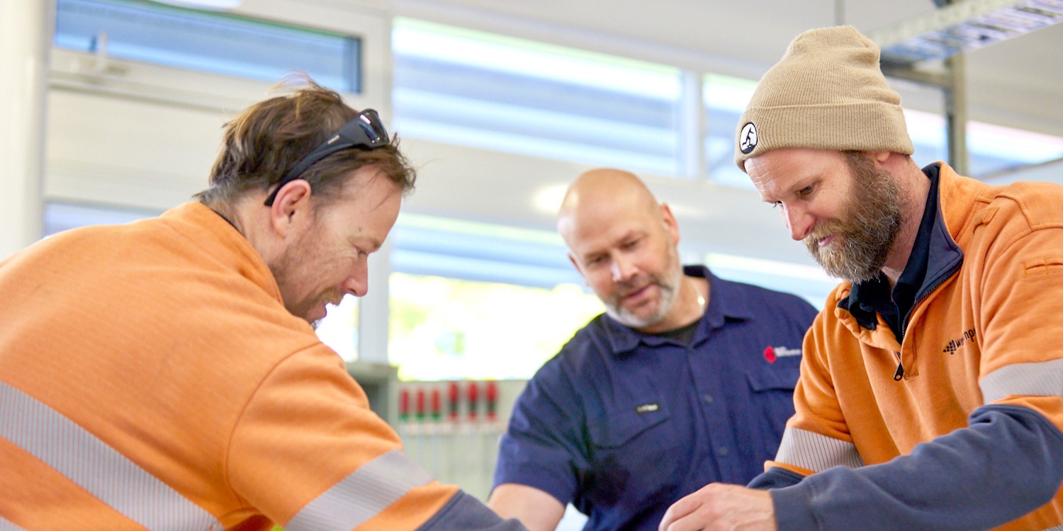 The scene captures three individuals engaging in a hands-on workshop. Two men in the foreground, dressed in casual work attire, are focused on assembling or adjusting components on a table. They are guided by a man in the background, possibly an instructor or supervisor. The room is well-lit with natural light coming through large windows, creating a collaborative and educational atmosphere.