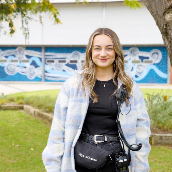 The image shows a young woman standing outdoors, smiling at the camera. She is wearing a light blue and white plaid jacket over a black top, with a utility belt or fanny pack labeled "Dept. of Justice." Attached to her jacket is a walkie-talkie, suggesting that she is involved in some official or security-related role. Behind her, there is a blue and white mural on a building, featuring abstract circular and wavy designs.